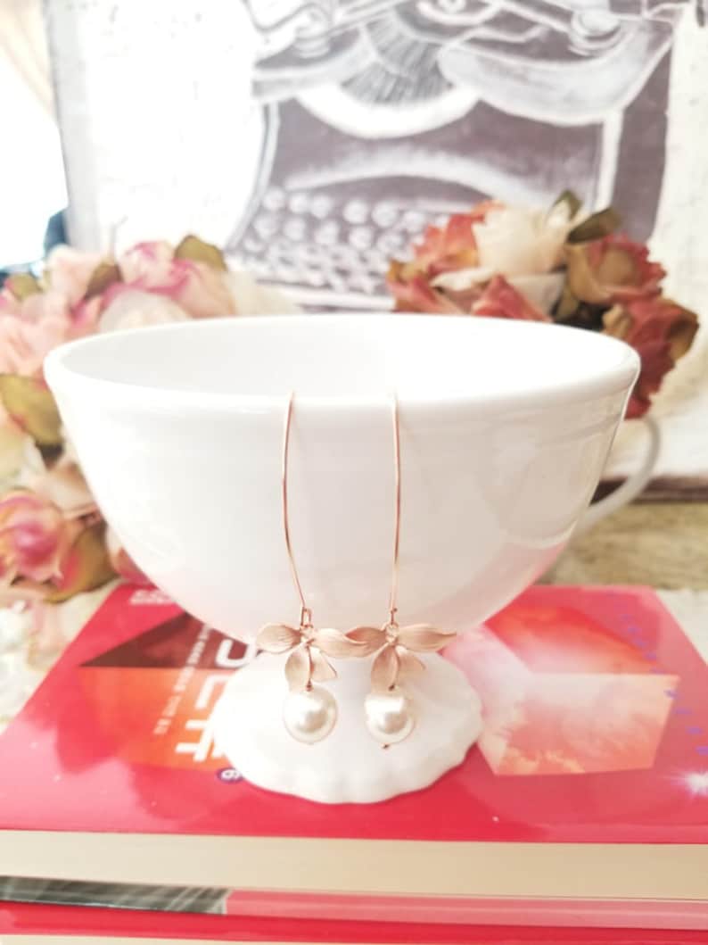 May include: A pair of delicate, rose gold earrings with a floral design and dangling faux pearls. The earrings are displayed on a white, seashell-shaped holder, set against a backdrop of a white bowl and a red book.