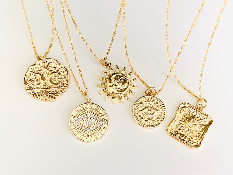 May include: Five gold necklaces with different pendants. The pendants are a sun and moon, an eye with a crystal, a square with a sun, and two different coins.