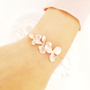 May include: A delicate silver bracelet featuring three flower-shaped charms. The bracelet is worn on a wrist, with the flowers arranged in a row. The flowers have a textured appearance and are connected by a fine chain.