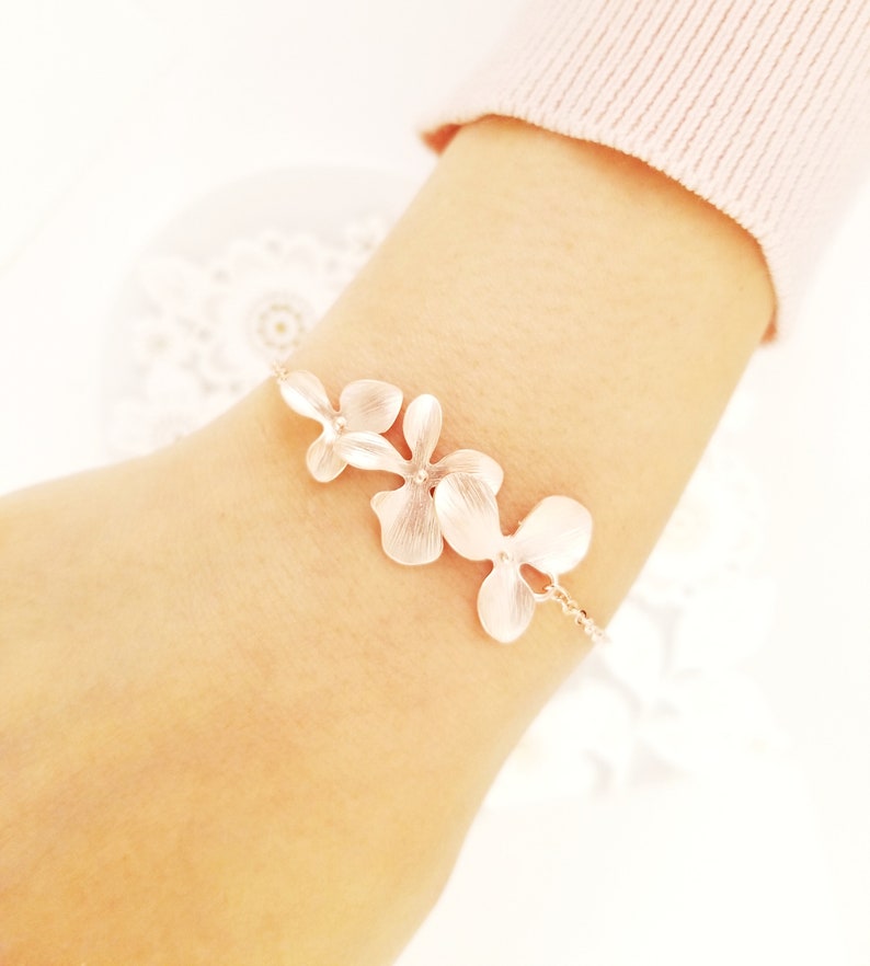 May include: A delicate silver bracelet featuring three flower-shaped charms. The bracelet is worn on a wrist, with the flowers arranged in a row. The flowers have a textured appearance and are connected by a fine chain.