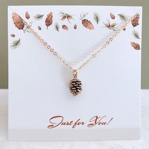 May include: A gold necklace with a small pine cone pendant. The necklace is displayed on a white card with the words "Just for You!" printed in brown.