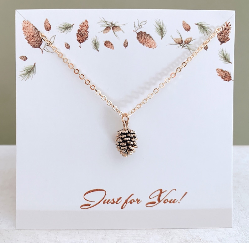 May include: A gold necklace with a small pine cone pendant. The necklace is displayed on a white card with the words "Just for You!" printed in brown.