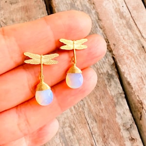 Op de afbeelding: Gouden oorhangers met libellenmotief en druppelvormige, opalescerende stenen. De libellenvleugels zijn gedetailleerd met geëtste lijnen en de stenen zijn gevat in gouden draad. De oorbellen worden getoond tegen een houten achtergrond.