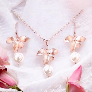 May include: A rose gold jewelry set featuring a necklace and earrings. The necklace and earrings have a floral design with a large pearl pendant. The jewelry is displayed on a white, textured surface with pink roses.