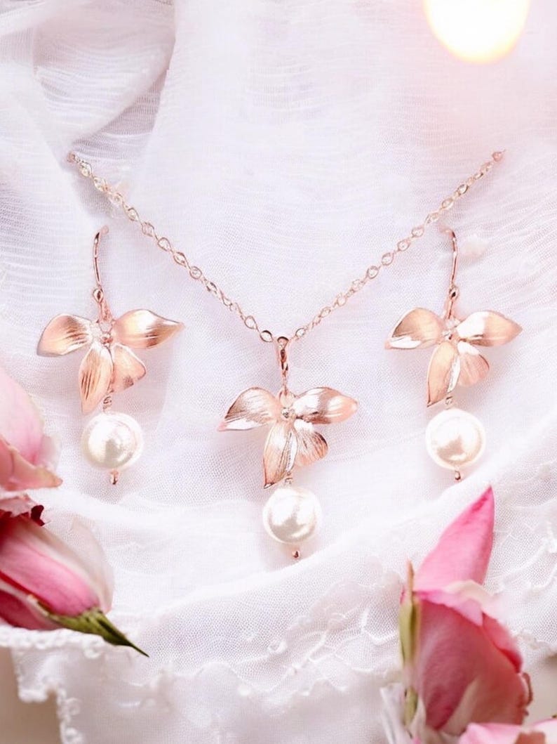 May include: A rose gold jewelry set featuring a necklace and earrings. The necklace and earrings have a floral design with a large pearl pendant. The jewelry is displayed on a white, textured surface with pink roses.