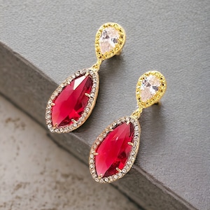 May include: A pair of gold earrings with red teardrop-shaped stones surrounded by clear crystals. The earrings have a smaller clear crystal at the top.