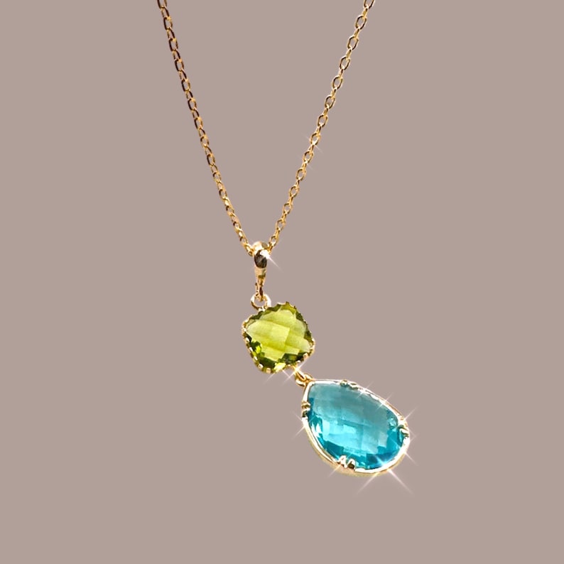 K&ouml;nnte beinhalten: Eine goldene Kettenkette mit einem Anh&auml;nger, der einen tropfenf&ouml;rmigen blauen Edelstein und einen kleineren quadratischen gr&uuml;nen Edelstein zeigt.
