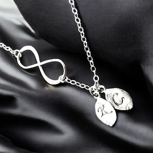 May include: A silver infinity necklace with two small silver charms. The charms are shaped like leaves and have the letters "K" and "S" engraved on them.