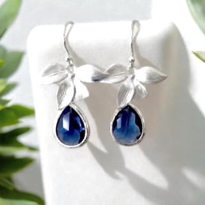 May include: A pair of silver-toned drop earrings. Each earring features a floral design at the top and a teardrop-shaped, deep blue gemstone. The earrings are displayed on a white surface, with green foliage in the background.