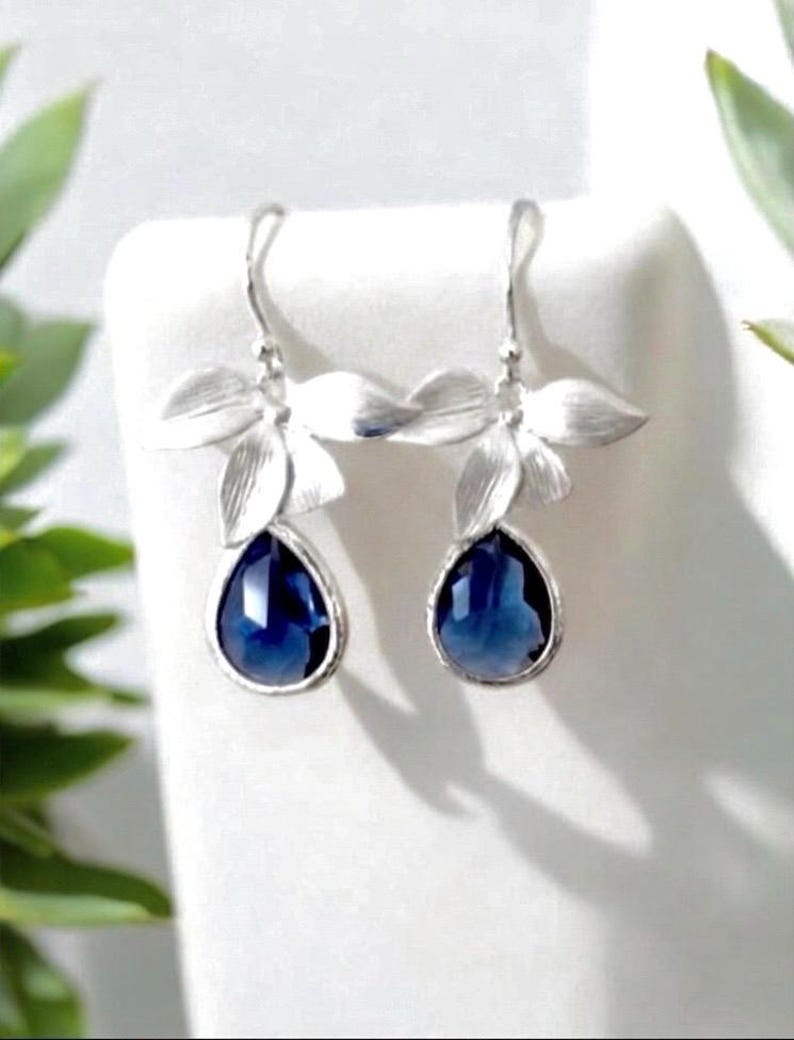 May include: A pair of silver-toned drop earrings. Each earring features a floral design at the top and a teardrop-shaped, deep blue gemstone. The earrings are displayed on a white surface, with green foliage in the background.