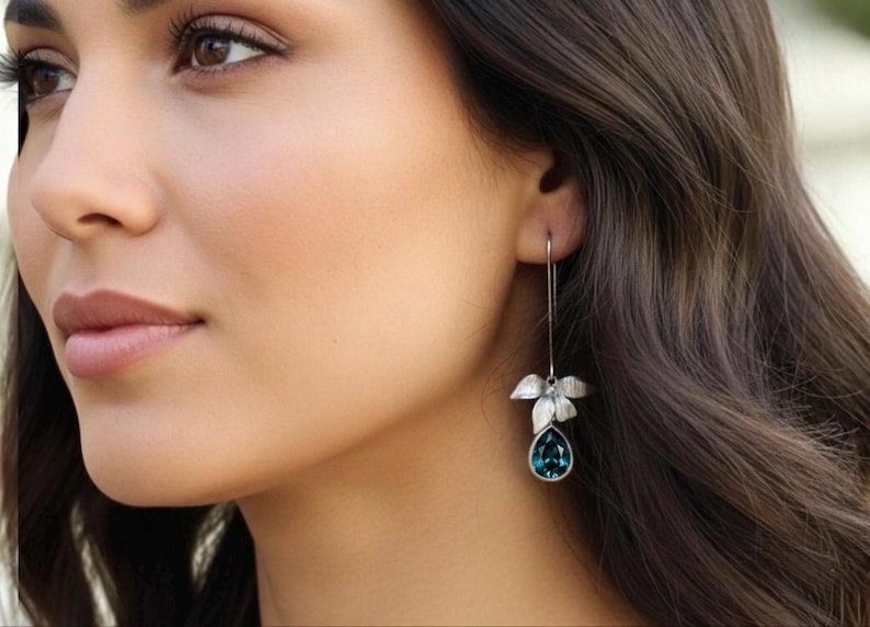 May include: A silver-toned earring with a leaf design and a teardrop-shaped teal gemstone. The earring hangs from a thin wire, showcasing a delicate and elegant style. The jewelry is a beautiful accessory.