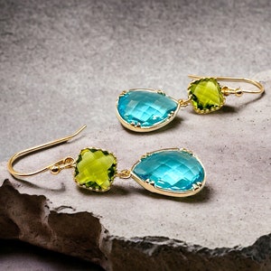 K&ouml;nnte beinhalten: Ein Paar goldene Ohrringe mit blauen und gr&uuml;nen tropfenf&ouml;rmigen Edelsteinen. Die Ohrringe haben ein h&auml;ngendes Design mit einem kleineren quadratischen Edelstein &uuml;ber dem gr&ouml;&szlig;eren Tropfen.