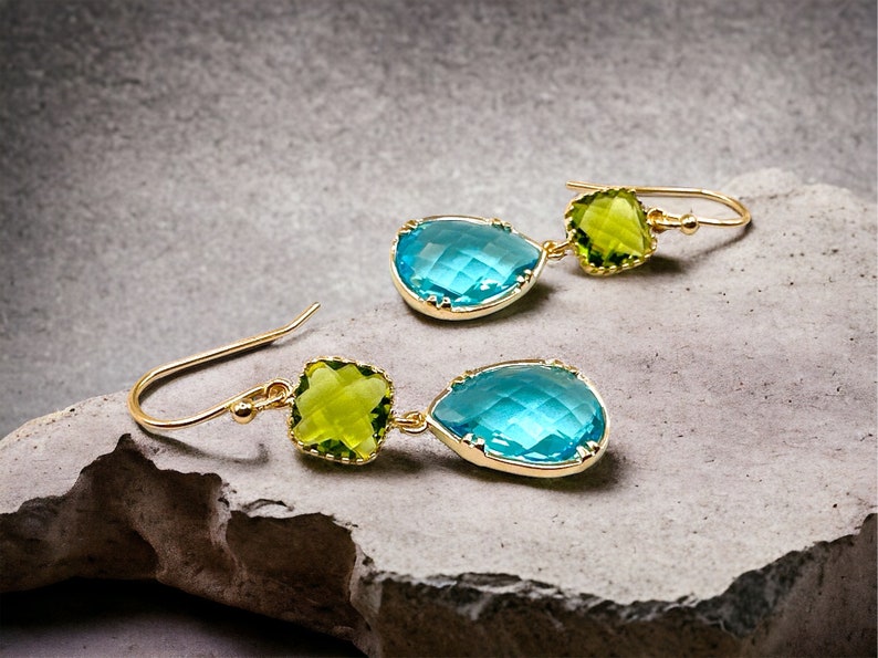 K&ouml;nnte beinhalten: Ein Paar goldene Ohrringe mit blauen und gr&uuml;nen tropfenf&ouml;rmigen Edelsteinen. Die Ohrringe haben ein h&auml;ngendes Design mit einem kleineren quadratischen Edelstein &uuml;ber dem gr&ouml;&szlig;eren Tropfen.