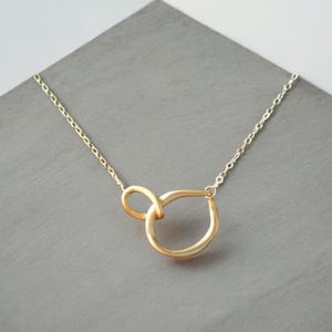 May include: A gold-colored necklace with a unique knot pendant. The pendant features a looped design, creating an abstract shape. The necklace chain is delicate and appears to be gold-toned. The jewelry is displayed on a gray surface.