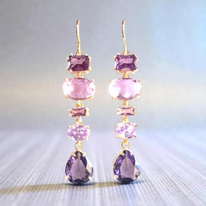Puede incluir: Un par de pendientes colgantes dorados con un degradado de piedras preciosas moradas. Cada pendiente tiene una piedra rectangular, ovalada, rectangular y en forma de lágrima. Las piedras hacen una transición de morado oscuro a claro.