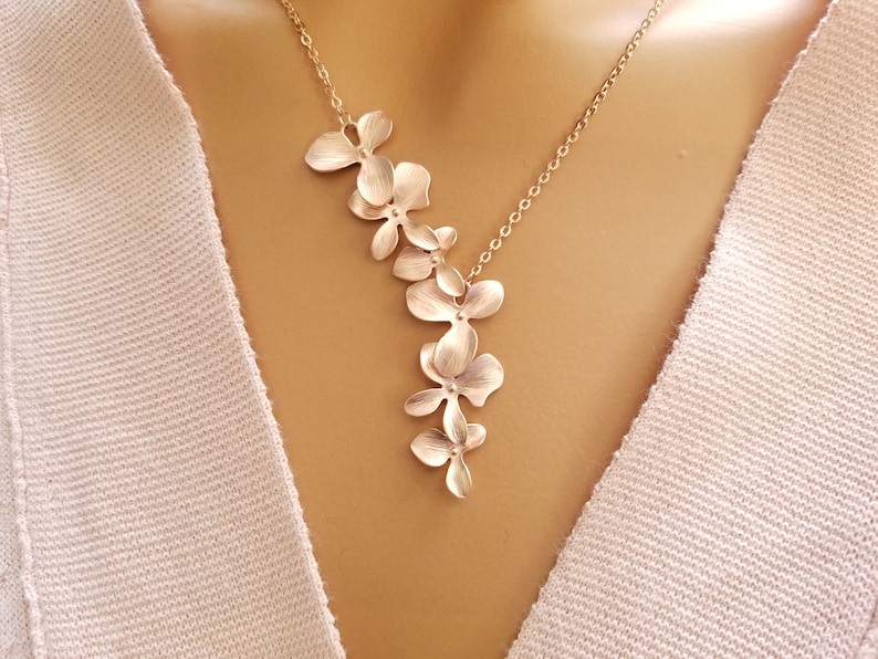 May include: A gold-colored necklace featuring a cascade of stylized flower pendants. The floral design is delicate and detailed, with each petal subtly textured. The necklace hangs on a fine chain, creating an elegant and feminine accessory.