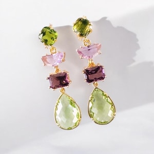 May include: A pair of gold-toned drop earrings featuring a series of colorful gemstones. The earrings showcase a combination of green, purple, and pink stones in various shapes, including round, rectangular, and teardrop. The earrings are designed to dangle.
