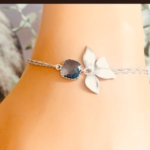 May include: Silver-toned anklet featuring a dark blue gemstone and a stylized flower. The anklet has a delicate, twisted chain design. The flower has four petals. The gemstone is a square shape with rounded edges.
