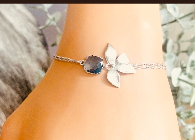 May include: Silver-toned anklet featuring a dark blue gemstone and a stylized flower. The anklet has a delicate, twisted chain design. The flower has four petals. The gemstone is a square shape with rounded edges.