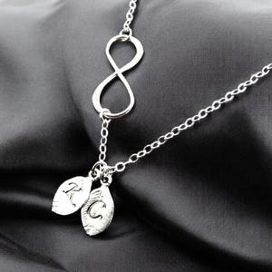 Könnte beinhalten: Eine silberne Unendlichkeitskette mit zwei kleinen Anhängern, die daran hängen. Die Anhänger sind blattförmig und haben die Buchstaben "K" und "C" eingraviert.