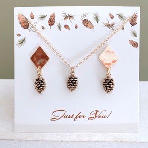 May include: A set of gold-tone earrings and a necklace featuring a pine cone charm. The earrings and necklace are displayed on a white card with a pine cone pattern and the text "Just for You!"