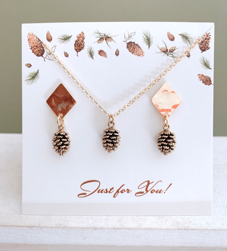 May include: A set of gold-tone earrings and a necklace featuring a pine cone charm. The earrings and necklace are displayed on a white card with a pine cone pattern and the text "Just for You!"