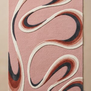 May include: A rectangular area rug featuring a dusty pink base with a raised, tufted abstract pattern of flowing, curved lines in shades of cream, terracotta orange, and charcoal grey. The design creates a layered, dimensional effect across the surface.