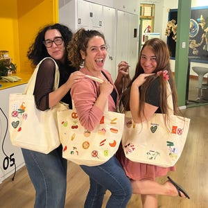 Peut inclure: Trois sacs fourre-tout en toile blanc cassé ornés de diverses icônes brodées colorées sur le thème de la nourriture et des vacances. Chaque sac présente des motifs uniques dont une pizza, un croissant, un hot-dog, des cerises et des palmiers.