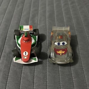 May include: Two toy race cars from the movie Cars. The left car is a red, white, and green Formula One style racer with the number 1. The right car is a translucent grey coupe with the number 95 on the roof and a red emblem on the hood.