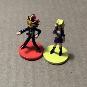 May include: Two small plastic figurines standing on circular bases. One figure has spiky black, red, and yellow hair, wearing a dark blue jacket and pants on a red base. The other figure has blonde hair, wearing a purple top and skirt on a yellow base.