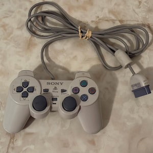 Könnte beinhalten: Ein hellgrauer, kabelgebundener Sony PSone-Gamecontroller mit zwei Analogsticks, einem Steuerkreuz, vier Aktionstasten mit geometrischen Symbolen, Select- und Start-Tasten sowie einer Analog-Taste, verbunden durch ein langes graues Kabel.
