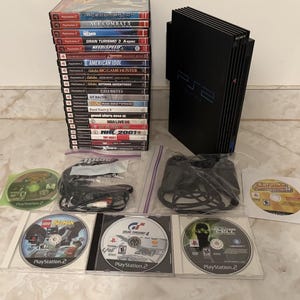 May include: A black Sony PlayStation 2 console standing vertically, accompanied by a stack of twenty PlayStation 2 game cases, a black wired controller, various loose game discs, and connection cables in clear plastic bags.
