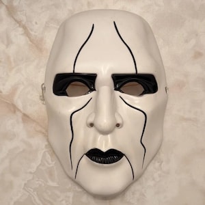 Op de afbeelding: Een wit volledig gezichtsmasker met opvallende zwarte beschilderde details. Het ontwerp bevat zwarte markeringen rond de oogkassen, verticale lijnen die vanaf het voorhoofd en de wangen lopen, en een zwart geschilderde mond met gedefinieerde lippen.