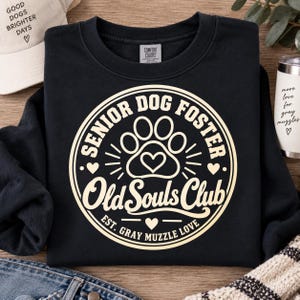 May include: A black crewneck sweatshirt featuring a cream-colored circular graphic. The design includes a paw print with a heart, the text "SENIOR DOG FOSTER Old Souls Club", and the phrase "EST. GRAY MUZZLE LOVE" with small heart accents.