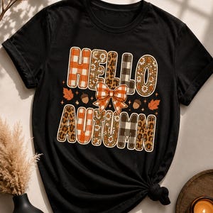 以下が含まれることがあります： 「HELLO AUTUMN」という文字が様々なパターンでデザインされた黒の半袖Tシャツ。文字にはオレンジのチェック柄、ヒョウ柄、白黒のチェック柄、花柄が使われています。中央にはオレンジのチェック柄のリボンが配置されています。