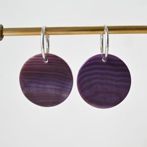 Op de afbeelding: Een paar oorringen met ronde, platte, paarskleurige hangers met horizontale, golvende, donkerdere paarse banden. De hangers hangen aan dunne, zilverkleurige metalen ringen die bevestigd zijn aan een horizontale goudkleurige stang.