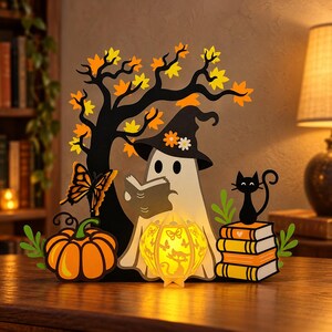 Puede incluir: Una figura decorativa de madera recortada que muestra un fantasma blanco con un sombrero de flores, leyendo un libro bajo un árbol negro con hojas naranjas y amarillas. Incluye una calabaza naranja, una pila de libros con un gato negro y una linterna brillante.
