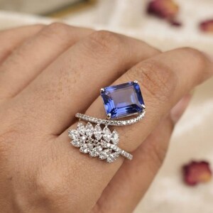 May include: A silver-toned ring featuring a large, rectangular, deep blue gemstone set horizontally. The band is open-style, with one side showcasing a curved row of small, clear, round stones and the other side featuring a cluster of marquise-cut stones.