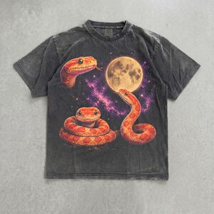 May include: A charcoal gray short-sleeve t-shirt featuring a graphic print of three orange snakes positioned around a large, glowing yellow full moon set against a backdrop of purple and black starry space, with a crew neckline and washed finish.