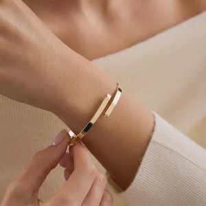 Könnte beinhalten: Ein goldfarbenes Metall-Armband in Form einer offenen Manschette mit flachen, polierten Oberflächen. Das Armband hat ein schlichtes, gewickeltes Design mit einer kleinen Lücke zwischen den beiden Enden und einer glatten, reflektierenden Oberfläche.