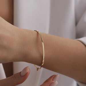 Peut inclure: Un bracelet jonc fin en or poli avec une surface lisse et réfléchissante. L'anneau circulaire présente un petit mécanisme de charnière intégré à la base, permettant de l'ouvrir et de le fermer solidement autour du poignet avec un fermoir.