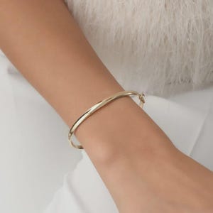 May include: A polished gold bangle bracelet with a smooth, rounded surface and a visible hinged clasp mechanism. The metal has a reflective, high-shine finish that catches the light, creating a clean and simple circular silhouette on the wrist.