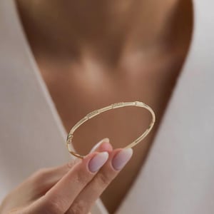 以下が含まれることがあります： 上半分に沿って小さな長方形のセグメントが並んだ、細いポリッシュ仕上げのゴールドバングルブレスレット。各セグメントには、小さく透明な輝く石が埋め込まれています。このブレスレットは、目に見える留め具が付いた繊細な円形デザインです。