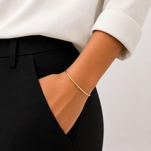 May include: A thin, polished gold bangle bracelet with a smooth, rounded surface. The bracelet is a simple, continuous circular band that rests on the wrist, showcasing a minimalist design with a reflective metallic finish and no additional embellishments.
