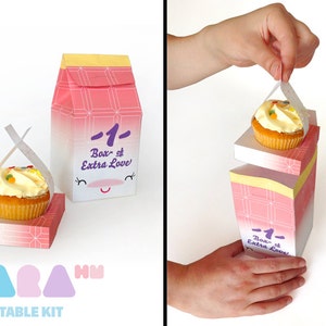 May include: A pink and white cardboard box with a smiling face and the text "1- Box of Extra Love" printed on it. The box is designed to hold a cupcake, which is shown being lifted out of the box. The box is part of a printable kit.
