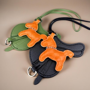 Op de afbeelding: Twee zadelvormige tashangers met oranje geweven paardfiguren. De ene hanger is olijfgroen met een bijpassende riem, de andere is zwart met een donkerblauwe riem. Elk paardfiguur is voorzien van een klein zilverkleurig kettinkje als detail.