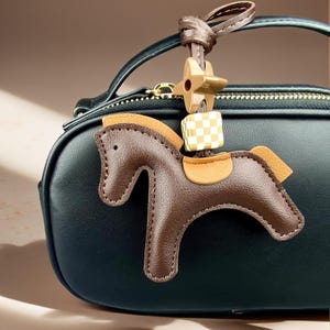 May include: A dark green handbag featuring a brown leather horse-shaped bag charm. The charm includes a tan saddle, a light brown mane, a small checkered cube bead, and a four-pointed star-shaped bead, all attached by a brown leather strap.