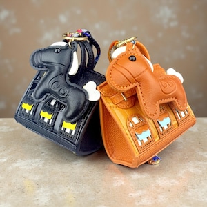 Op de afbeelding: Twee miniatuur handtasbedels, één zwart en één oranje, elk voorzien van een gestikt paardvormig lederen ornament bevestigd aan een goudkleurige metalen ring. De tassen hebben gestructureerde oppervlakken, kleine handvatten en uitsparingen.