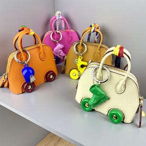May include: Four miniature dome-shaped handbags in orange, pink, mustard yellow, and cream, each featuring two top handles, a zipper closure, and a roller skate keychain charm. The bags have two small wheels attached to the bottom front corners.