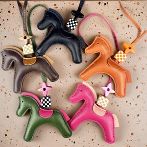 May include: Five horse-shaped bag charms made of leather in black, brown, orange, olive green, and magenta. Each charm features a saddle, a small checkered cube bead, and a star-shaped connector attached to a thin leather cord for hanging.
